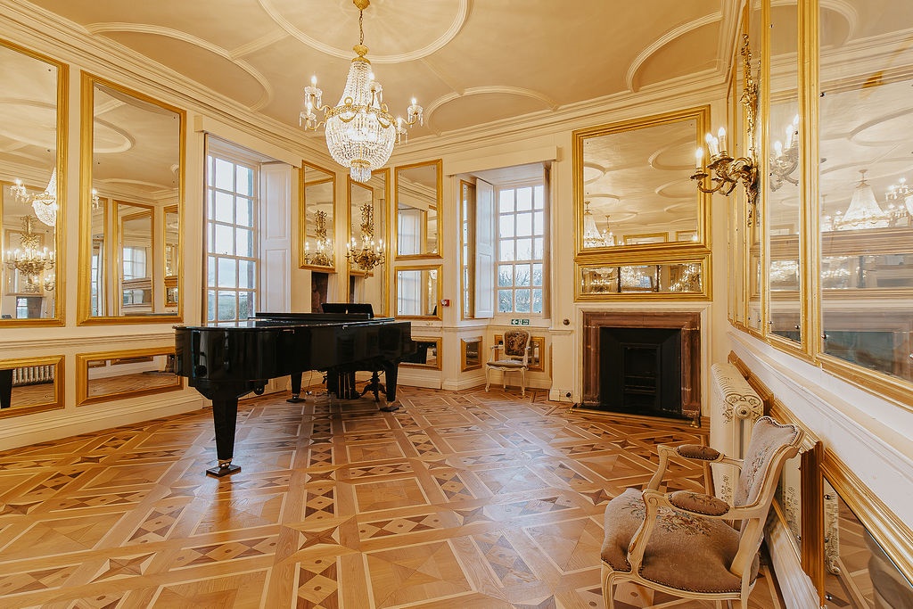An opulent hall featuring intricate parquet wood flooring and walls adorned with large, gold-framed mirrors and elegant wall sconces. A black grand piano sits near tall windows, while a crystal chandelier hangs from the ornate white ceiling. The room also includes a marble fireplace and classic upholstered chairs.