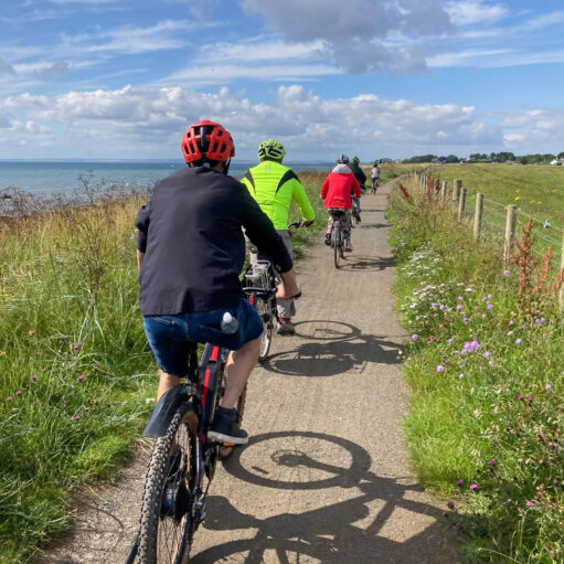 A view from behind a group of cyclists riding in a line along a narrow dirt path. To their left is a grassy bank leading down to the sea, and to their right is a wooden fence bordering a green field under a partly cloudy blue sky.