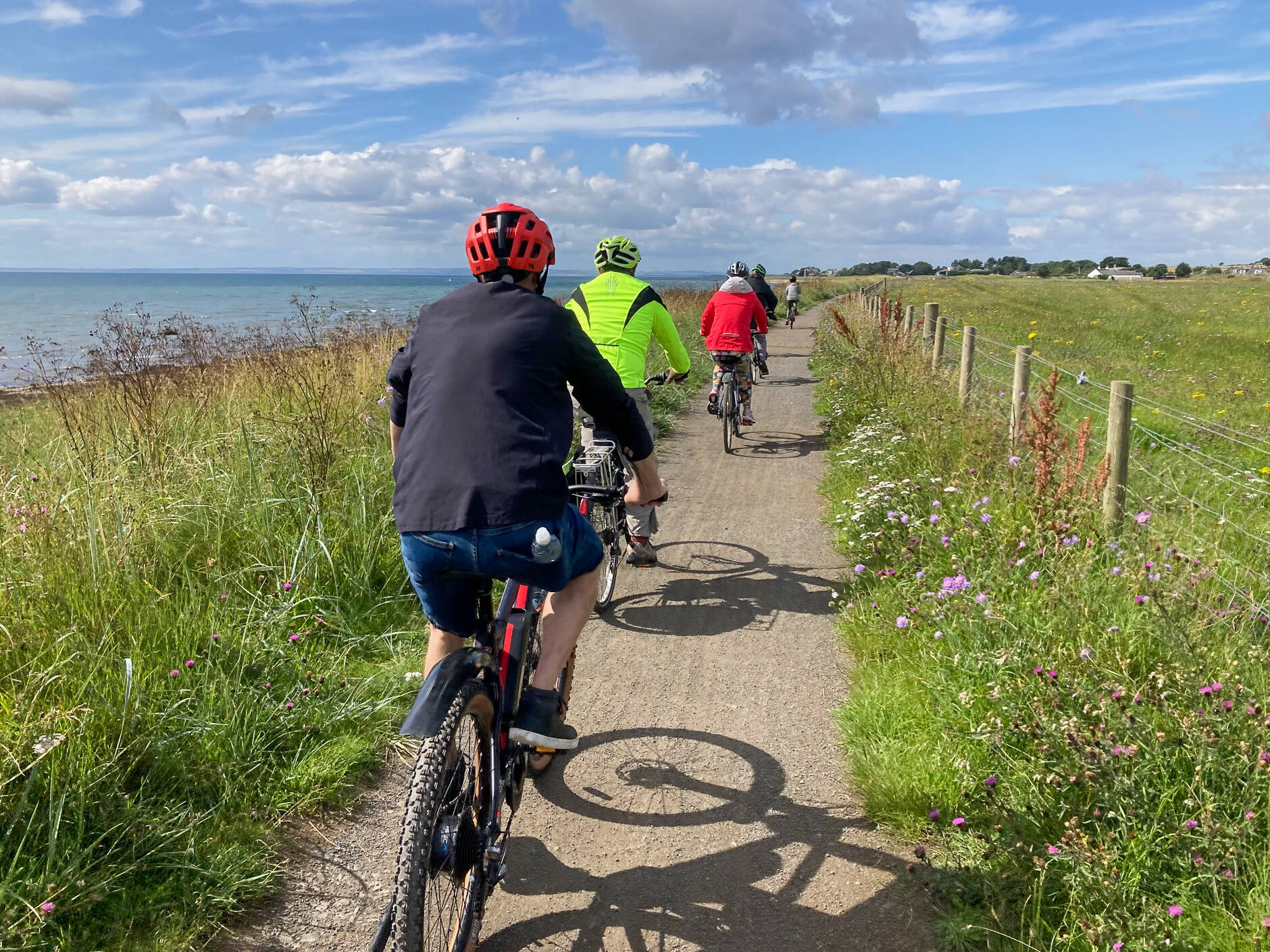 A view from behind a group of cyclists riding in a line along a narrow dirt path. To their left is a grassy bank leading down to the sea, and to their right is a wooden fence bordering a green field under a partly cloudy blue sky.