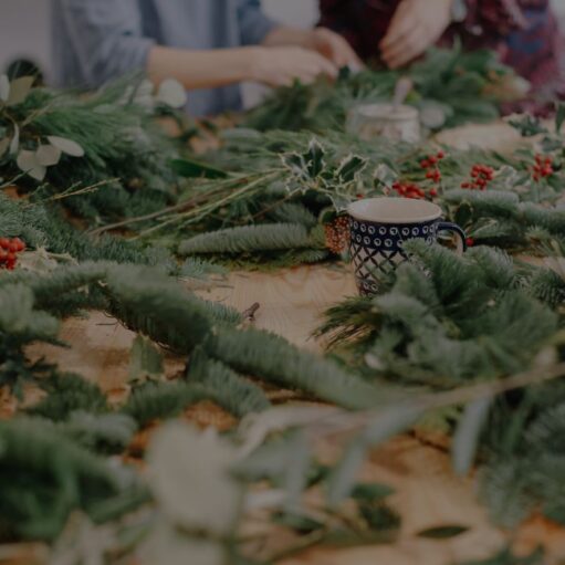 A close-up, slightly high-angle shot of a wooden table covered in festive foliage, including pine branches, holly, and red berries. A patterned ceramic mug sits amidst the greenery, while the blurred hands of people assembling winter wreaths are visible in the background.