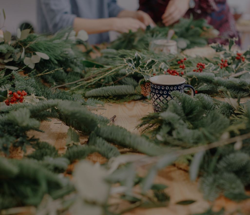 A close-up, slightly high-angle shot of a wooden table covered in festive foliage, including pine branches, holly, and red berries. A patterned ceramic mug sits amidst the greenery, while the blurred hands of people assembling winter wreaths are visible in the background.