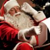 A close-up shot of Santa Claus wearing his classic red suit, hat, and white gloves, adjusting his glasses with one hand while holding up a long list of paper to read. A decorated Christmas tree with a silver ornament is visible in the background.