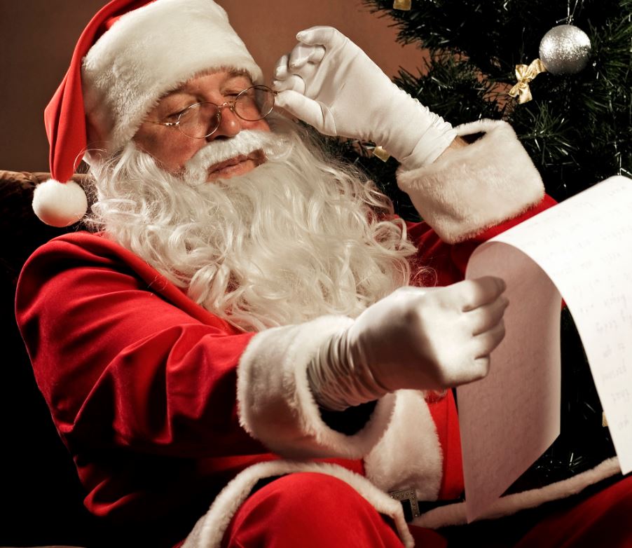 A close-up shot of Santa Claus wearing his classic red suit, hat, and white gloves, adjusting his glasses with one hand while holding up a long list of paper to read. A decorated Christmas tree with a silver ornament is visible in the background.