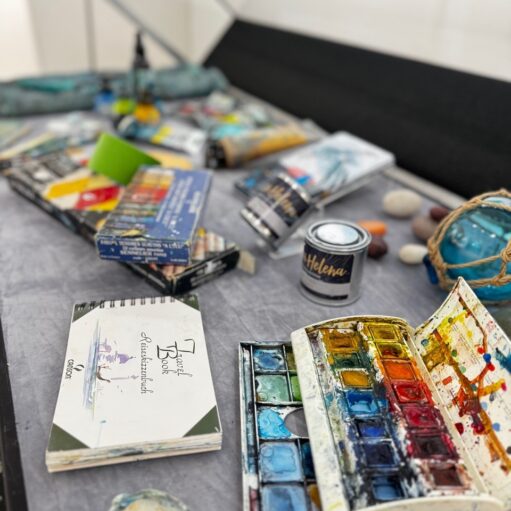 An assortment of watercolor painting supplies arranged on a grey surface, including a spiral-bound travel sketchbook, an open watercolor palette with various paint wells, paint tubes, boxes of Sennelier pastels, and small cans of "Helena" paint.