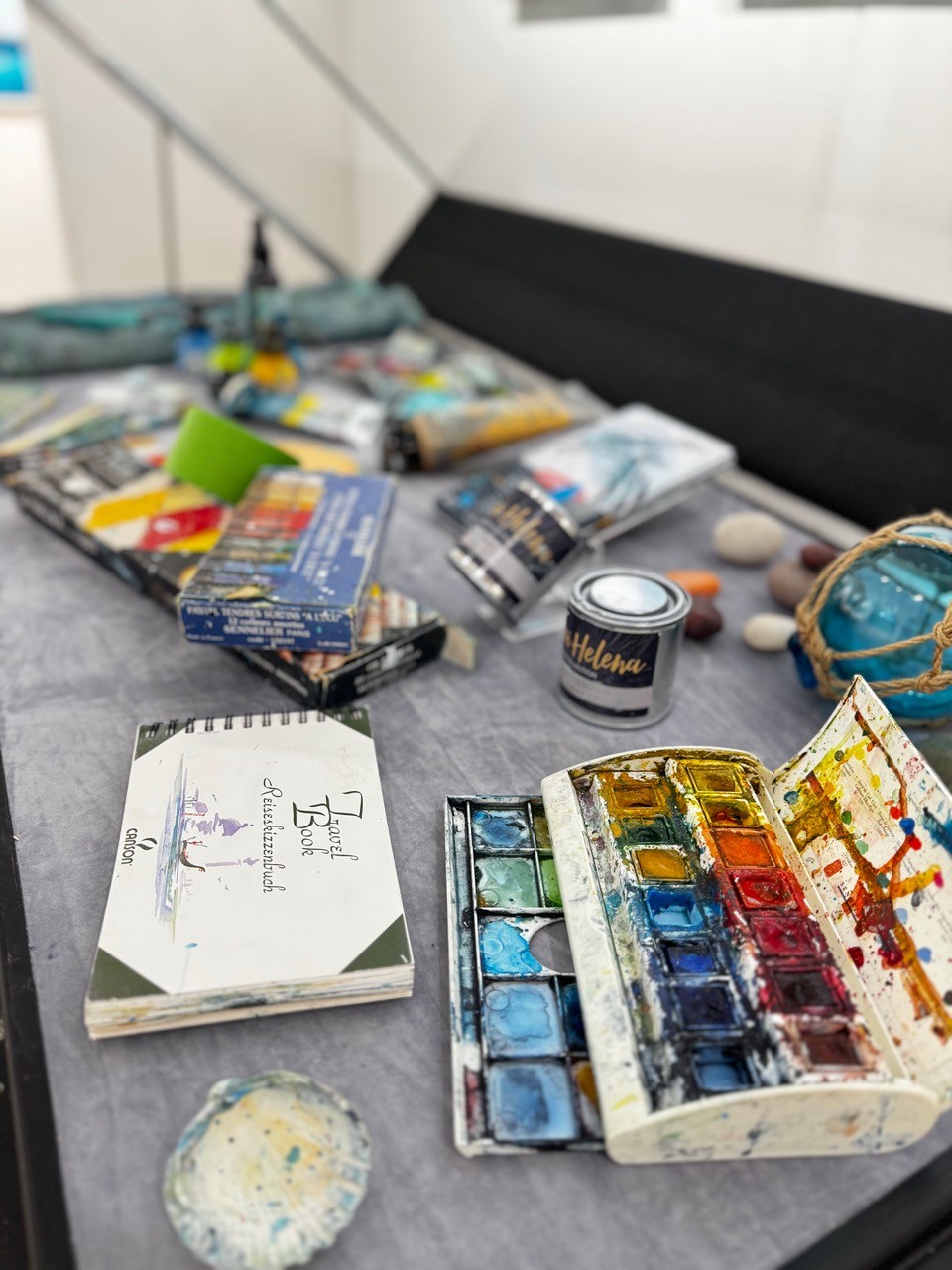An assortment of watercolor painting supplies arranged on a grey surface, including a spiral-bound travel sketchbook, an open watercolor palette with various paint wells, paint tubes, boxes of Sennelier pastels, and small cans of "Helena" paint.