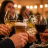 A close-up shot showing a group of people raising their glasses together in a celebratory toast. The selection of alcoholic drinks includes a glass of white wine, a pint of foamy beer, a cocktail garnished with a lemon slice and a sprig of rosemary, and a glass of red wine. The background is softly blurred with warm, glowing string lights and indistinct figures at a social gathering or party.
