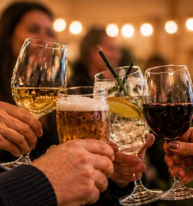 A close-up shot showing a group of people raising their glasses together in a celebratory toast. The selection of alcoholic drinks includes a glass of white wine, a pint of foamy beer, a cocktail garnished with a lemon slice and a sprig of rosemary, and a glass of red wine. The background is softly blurred with warm, glowing string lights and indistinct figures at a social gathering or party.