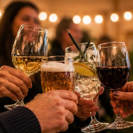 A close-up shot showing a group of people raising their glasses together in a celebratory toast. The selection of alcoholic drinks includes a glass of white wine, a pint of foamy beer, a cocktail garnished with a lemon slice and a sprig of rosemary, and a glass of red wine. The background is softly blurred with warm, glowing string lights and indistinct figures at a social gathering or party.