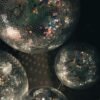 A low-angle view featuring a cluster of multiple mirrored disco balls of varying sizes suspended against a dark background. The tiny square mirrors on each sphere catch and reflect soft, colorful light, casting subtle glimmers and reflections across the surfaces.