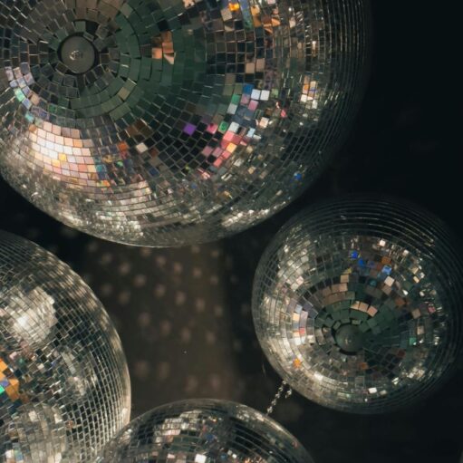 A low-angle view featuring a cluster of multiple mirrored disco balls of varying sizes suspended against a dark background. The tiny square mirrors on each sphere catch and reflect soft, colorful light, casting subtle glimmers and reflections across the surfaces.