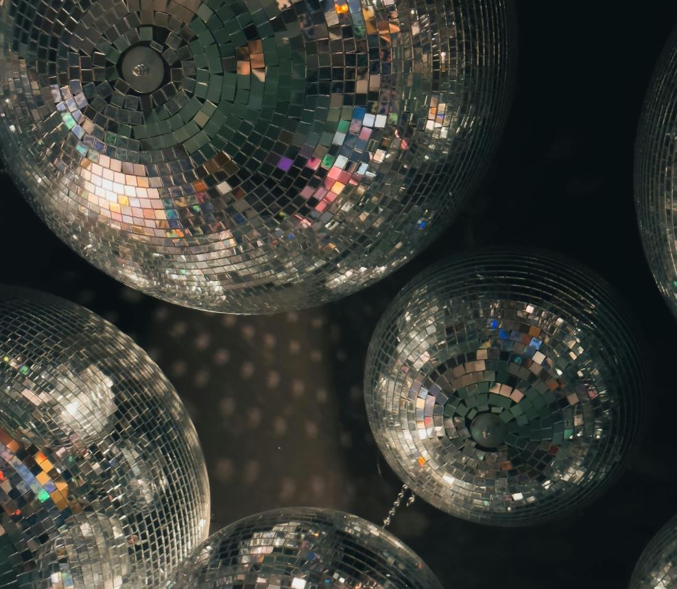 A low-angle view featuring a cluster of multiple mirrored disco balls of varying sizes suspended against a dark background. The tiny square mirrors on each sphere catch and reflect soft, colorful light, casting subtle glimmers and reflections across the surfaces.