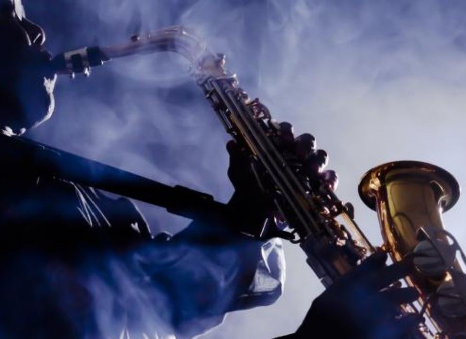 A musician playing a brass saxophone. The musician is silhouetted against a bright, hazy light source, with thick plumes of blue-tinted smoke or fog swirling around them and the instrument, creating an atmospheric, moody jazz club vibe.