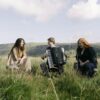 Three musicians sitting in a tall, grassy field with rolling green hills and a bright sky in the background. In the center, a young man plays a large black accordion, smiling toward a woman on the left who wears a long patterned dress. On the right, a woman with long red hair wearing a dark dress laughs, enjoying the performance.