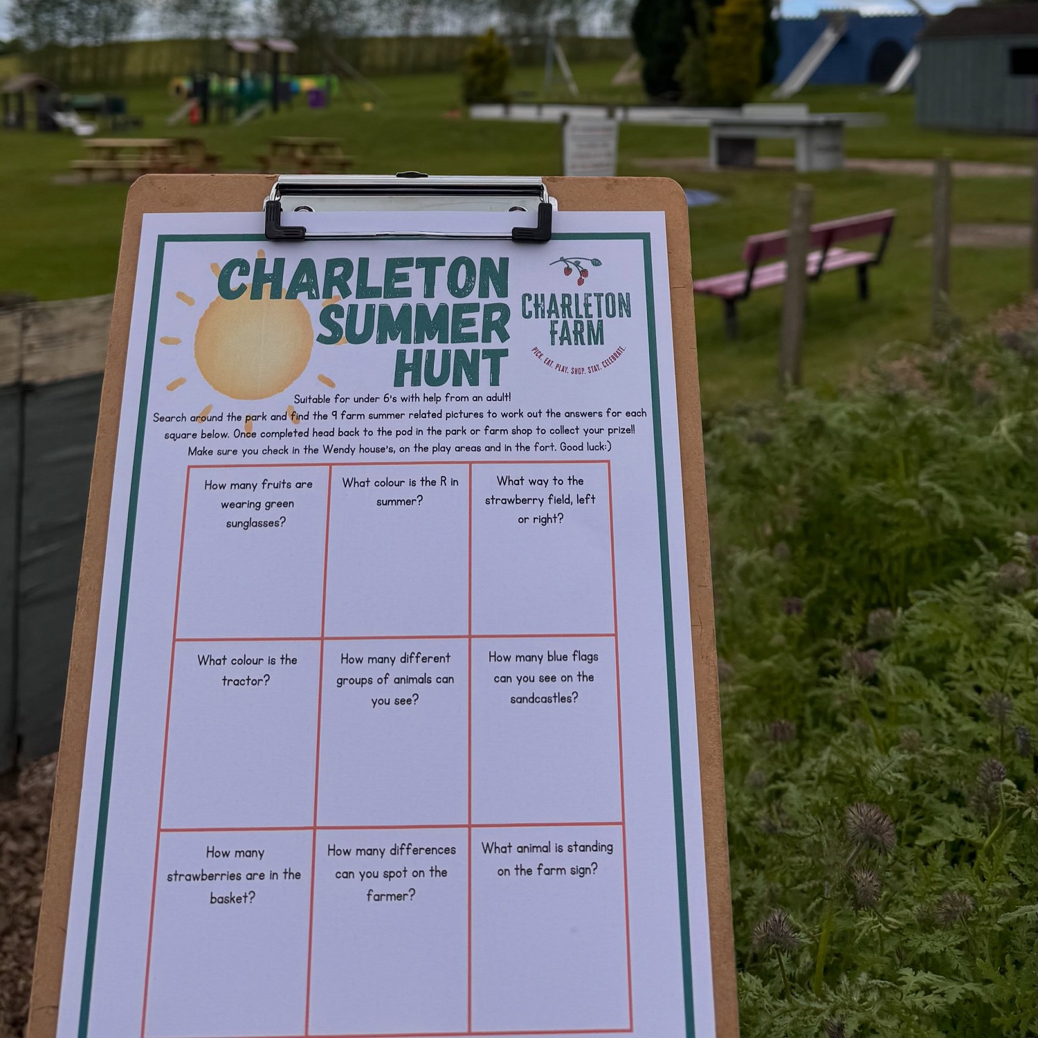 Clipboard displaying a “Charleton Summer Hunt” activity sheet in a farm park, with playground equipment, benches, and grassy outdoor areas visible in the background.