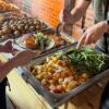 Self-service carvery buffet with trays of roast vegetables, potatoes, and gravy as a person plates a meal with meat, Yorkshire pudding, and green beans.