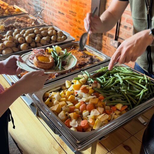 Self-service carvery buffet with trays of roast vegetables, potatoes, and gravy as a person plates a meal with meat, Yorkshire pudding, and green beans.