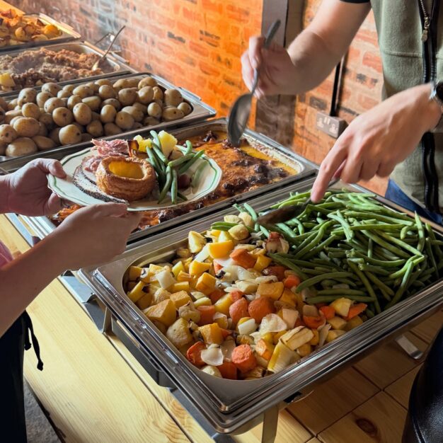 Self-service carvery buffet with trays of roast vegetables, potatoes, and gravy as a person plates a meal with meat, Yorkshire pudding, and green beans.