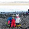 Two children in bright waterproof clothing stand on a rocky shoreline with an adult, exploring tidal pools beside a calm estuary.