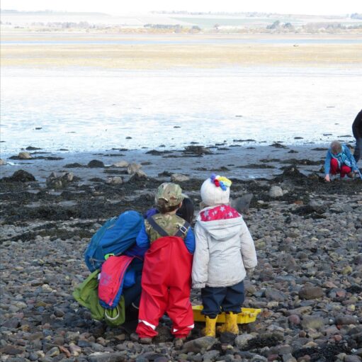 Two children in bright waterproof clothing stand on a rocky shoreline with an adult, exploring tidal pools beside a calm estuary.