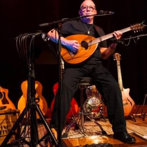 Performer seated on stage playing a mandolin, surrounded by guitars and microphones during a live music setup.