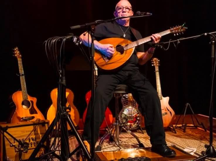 Performer seated on stage playing a mandolin, surrounded by guitars and microphones during a live music setup.
