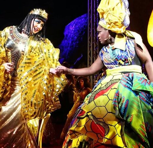 Stage performance with two costumed actors in elaborate, brightly coloured outfits—one in gold and the other in patterned fabrics—reaching toward each other under theatrical lighting.