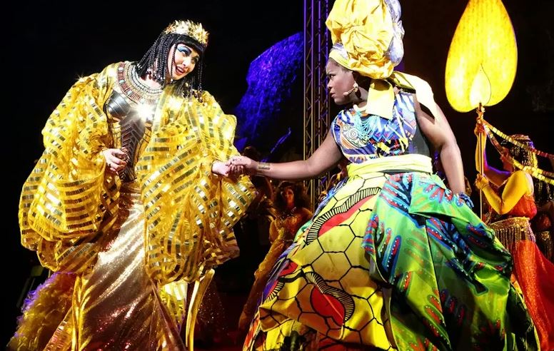 Stage performance with two costumed actors in elaborate, brightly coloured outfits—one in gold and the other in patterned fabrics—reaching toward each other under theatrical lighting.