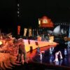 Large-scale night performance featuring a giant dragon head prop, costumed performers on a lit stage, and tall decorative set pieces with a waterfront cityscape in the background.