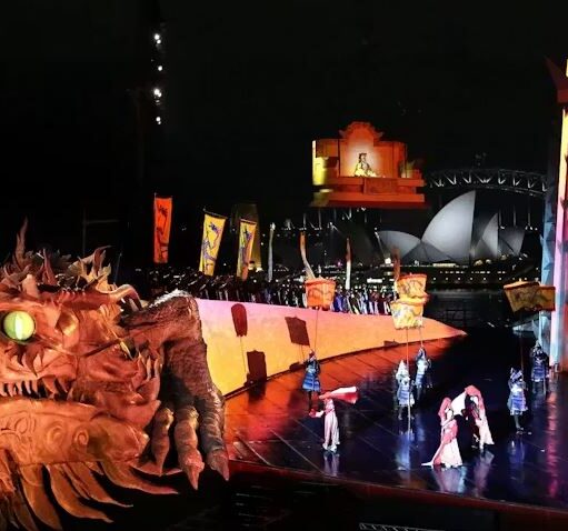 Large-scale night performance featuring a giant dragon head prop, costumed performers on a lit stage, and tall decorative set pieces with a waterfront cityscape in the background.