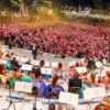 Large outdoor concert with an orchestra on stage and a huge crowd gathered in front, confetti falling over the audience.