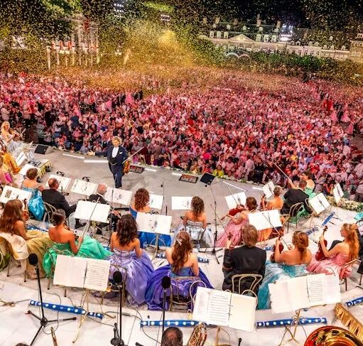 Large outdoor concert with an orchestra on stage and a huge crowd gathered in front, confetti falling over the audience.
