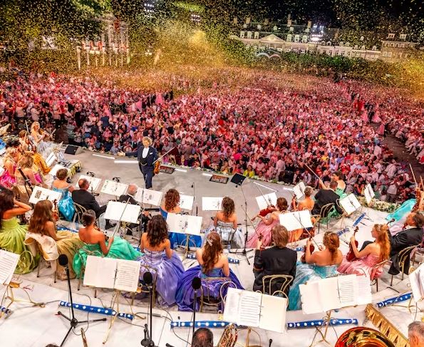 Large outdoor concert with an orchestra on stage and a huge crowd gathered in front, confetti falling over the audience.