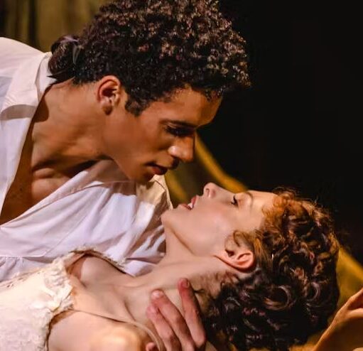 Two actors in a stage scene, one supporting the other in a dramatic dip as they lean close under warm theatrical lighting.
