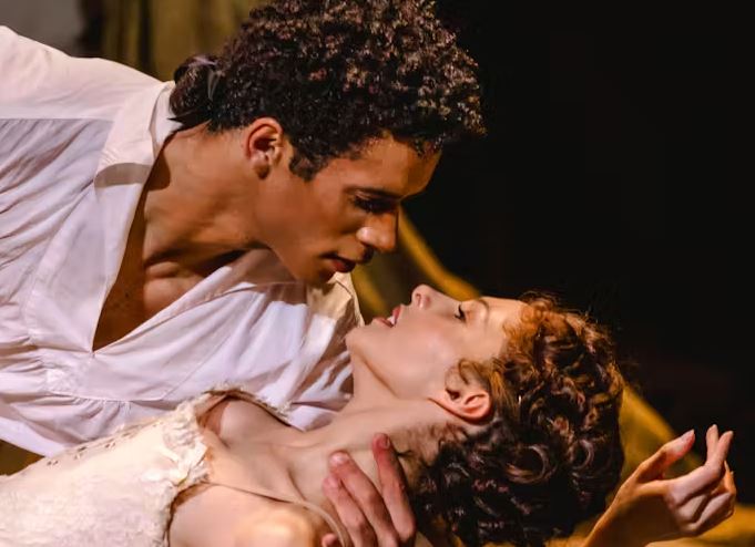 Two actors in a stage scene, one supporting the other in a dramatic dip as they lean close under warm theatrical lighting.