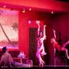 Stage performance in a red-lit lounge setting, with three actors posing mid-dance on and around chairs and a sofa, drinks set on side tables.