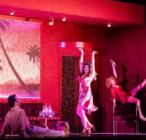 Stage performance in a red-lit lounge setting, with three actors posing mid-dance on and around chairs and a sofa, drinks set on side tables.