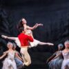 Ballet performance on stage with a dancer in a white costume lifted by a partner in a red jacket, surrounded by an ensemble in matching white outfits against a woodland backdrop.