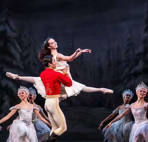 Ballet performance on stage with a dancer in a white costume lifted by a partner in a red jacket, surrounded by an ensemble in matching white outfits against a woodland backdrop.