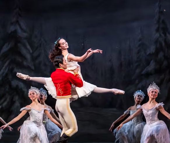 Ballet performance on stage with a dancer in a white costume lifted by a partner in a red jacket, surrounded by an ensemble in matching white outfits against a woodland backdrop.