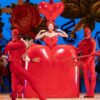Stage scene with performers in striking red costumes, including a central figure inside a large heart-shaped prop, surrounded by others holding props against a colourful backdrop.
