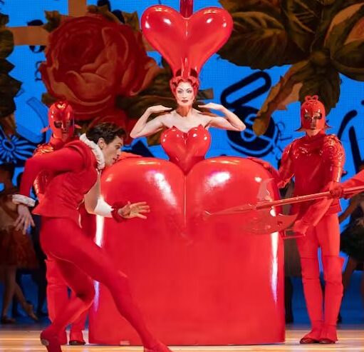 Stage scene with performers in striking red costumes, including a central figure inside a large heart-shaped prop, surrounded by others holding props against a colourful backdrop.