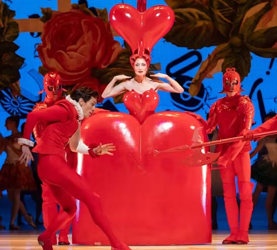 Stage scene with performers in striking red costumes, including a central figure inside a large heart-shaped prop, surrounded by others holding props against a colourful backdrop.
