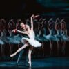 Ballet scene with a dancer in a white tutu posed en pointe, supported by a partner, with a line of similarly dressed dancers in the background.