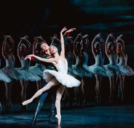 Ballet scene with a dancer in a white tutu posed en pointe, supported by a partner, with a line of similarly dressed dancers in the background.