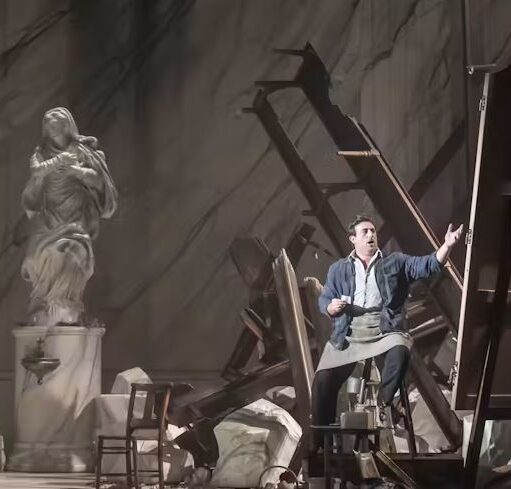 Stage scene depicting a workshop-like setting, with a performer reaching outward among scattered wooden frames, broken pieces, and a classical statue on a pedestal.