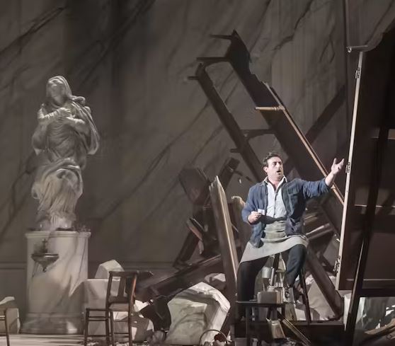Stage scene depicting a workshop-like setting, with a performer reaching outward among scattered wooden frames, broken pieces, and a classical statue on a pedestal.
