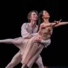 Two ballet dancers in flowing costumes perform a lift on stage, with one extending an arm outward while balanced in the partner’s arms against a dark background.