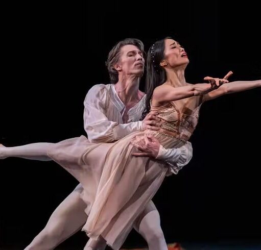 Two ballet dancers in flowing costumes perform a lift on stage, with one extending an arm outward while balanced in the partner’s arms against a dark background.