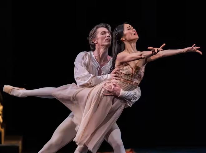 Two ballet dancers in flowing costumes perform a lift on stage, with one extending an arm outward while balanced in the partner’s arms against a dark background.