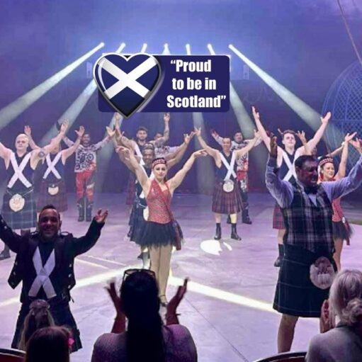 Stage performance with kilted dancers raising their arms in unison, a Scottish flag emblem and the text “Proud to be in Scotland” displayed above, while audience members watch and applaud in the foreground.